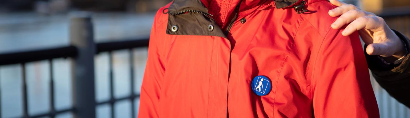 etualalla henkilö, jolla on takissa näkövammaismerkki ja hänen olkapäällä on avustajan käsi